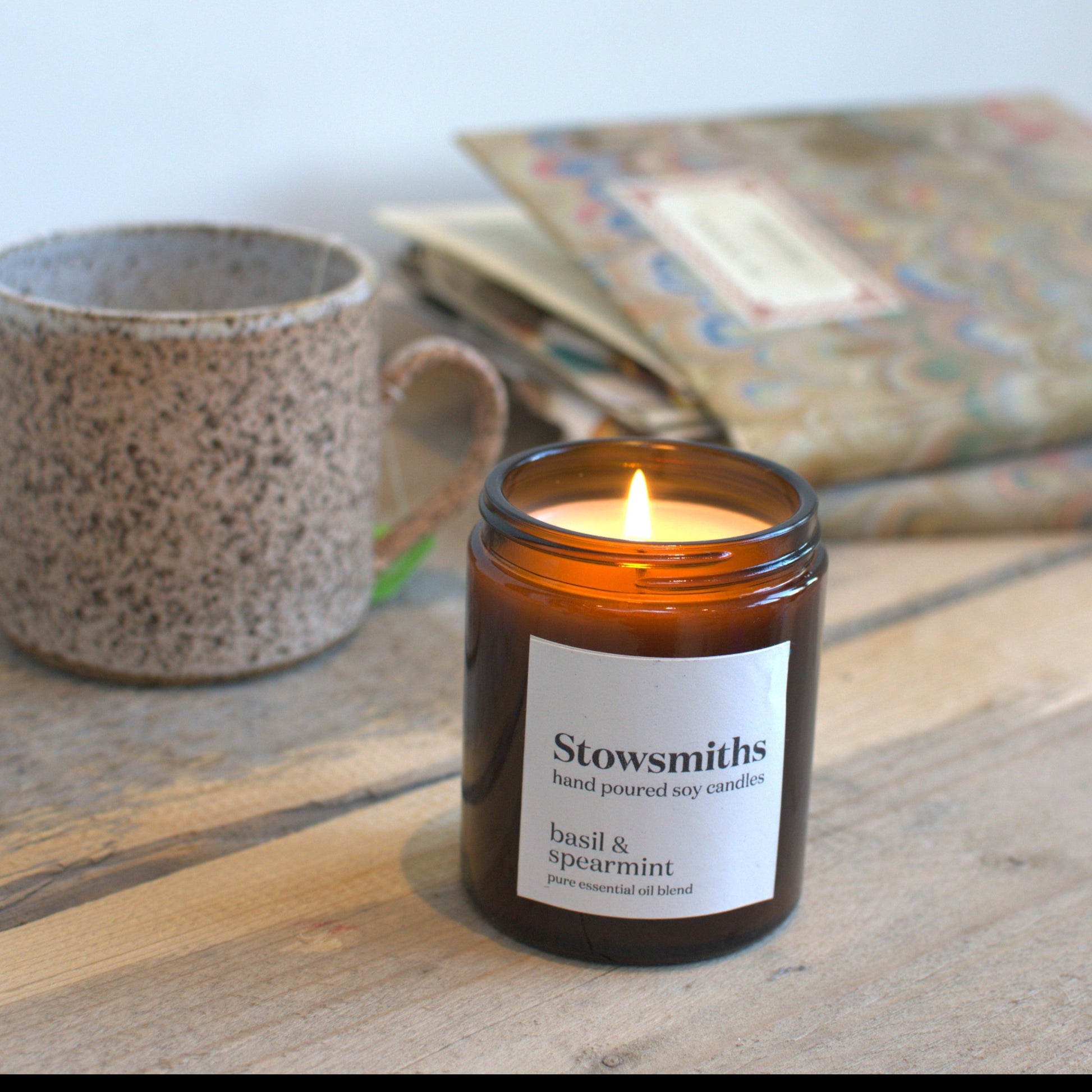 Candle in a glass jar labeled 'Stowsmiths' on a wooden surface with a speckled mug and patterned fabric in the background.